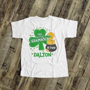 K&ouml;nnte beinhalten: Wei&szlig;es T-Shirt mit einem gr&uuml;nen Kleeblatt und dem Text "This Little Shamrock is Two!" mit einem Topf voller Gold und dem Namen "Dalton" darauf gedruckt.