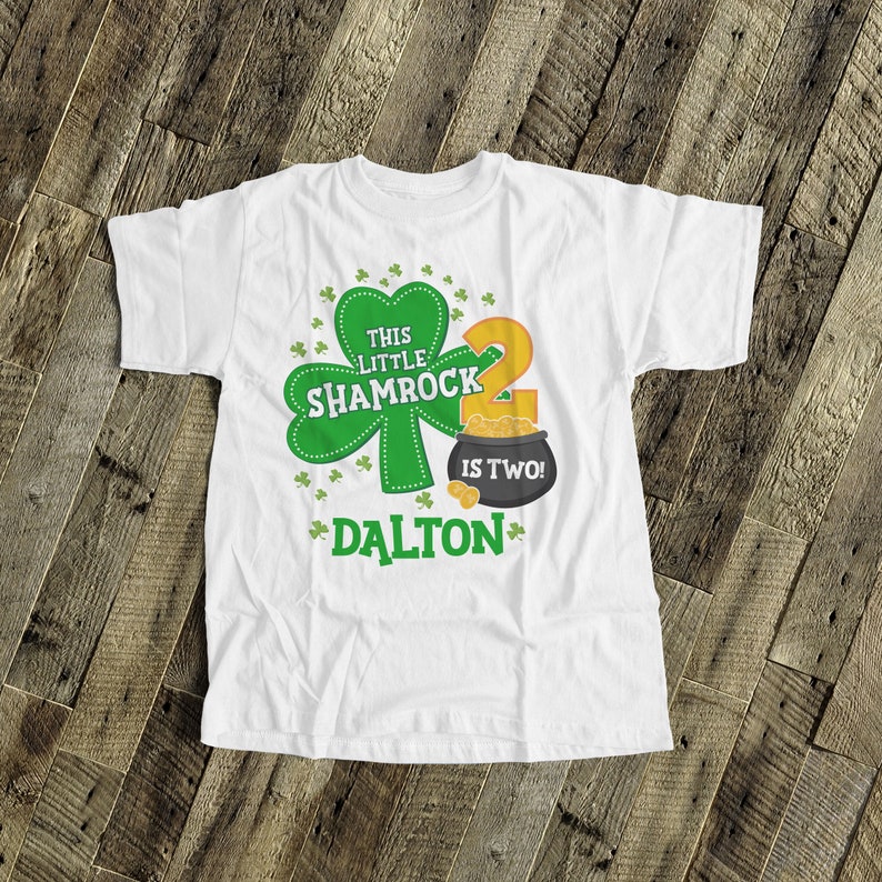 K&ouml;nnte beinhalten: Wei&szlig;es T-Shirt mit einem gr&uuml;nen Kleeblatt und dem Text "This Little Shamrock is Two!" mit einem Topf voller Gold und dem Namen "Dalton" darauf gedruckt.