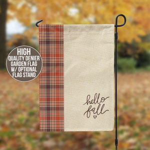 May include: A rectangular garden flag with a plaid pattern on the left side in shades of brown, orange, and red. The right side is a beige color with the words "hello fall" in a cursive font. A black metal flag stand is on the right.