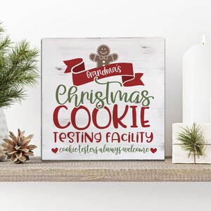 May include: A white wooden sign with red and green text that reads "Grandma's Christmas Cookie Testing Facility" with a heart on either side of the text "cookie testers always welcome". A gingerbread man cookie with a bow tie is above the text "Grandma's".