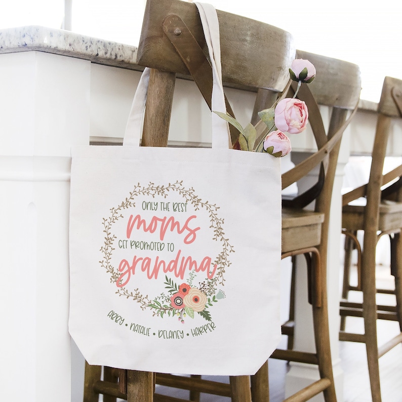 May include: A white canvas tote bag featuring the text "Only the best moms get promoted to grandma" within a floral wreath design. The names "Abby, Natalie, Delaney, Harper" are listed below the wreath.