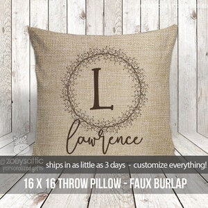 May include: A 16x16 inch throw pillow with a burlap texture. The pillow features a brown wreath with a large letter "L" in the center and the name "lawrence" written below the wreath.