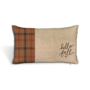 May include: Rectangular decorative pillow with a plaid pattern in shades of brown and orange on the left side. The right side is a tan burlap texture with the words "hello fall" in a handwritten font.