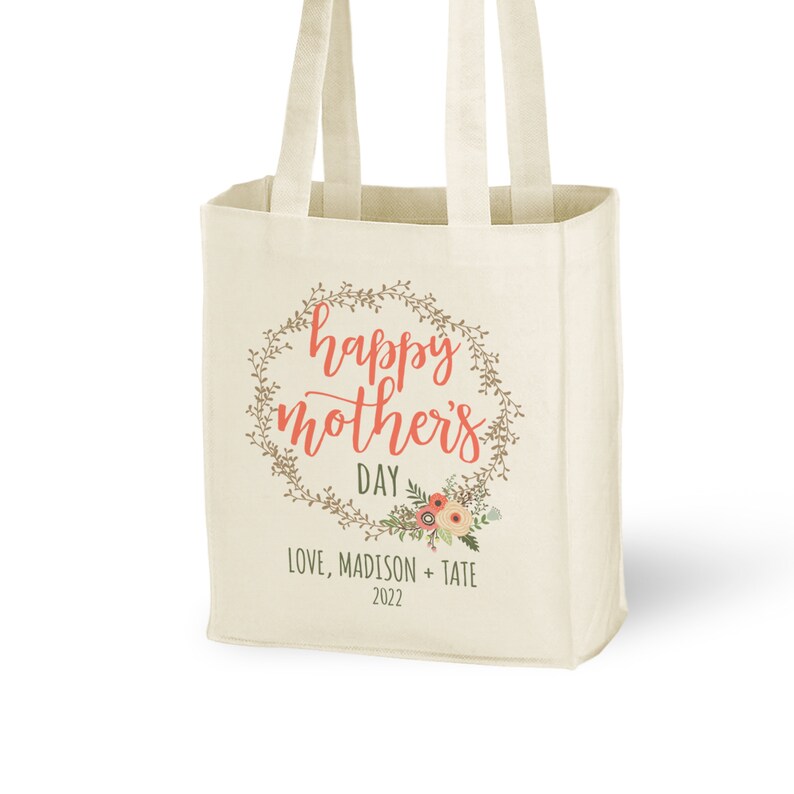 May include: A white canvas tote bag with a floral wreath design and the text "happy mother's day" in coral pink lettering. The text "LOVE, MADISON + TATE 2022" is printed below the wreath.