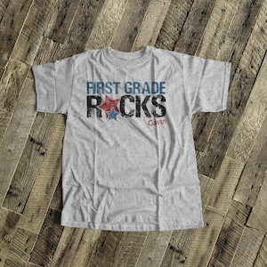 May include: A gray t-shirt with the text "First Grade Rocks" in black with red and blue stars. The name "Gavin" is printed below the text.