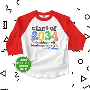 May include: White and red baseball style t-shirt with the text "class of 2034 kicking it off kindergarten style -Caden" in colorful handwritten font.