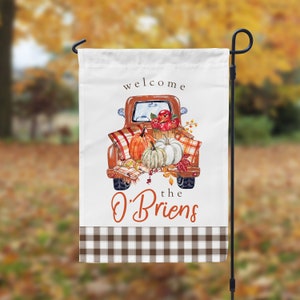 Peut inclure: Bienvenue chez les O'Briens ! Un drapeau de jardin blanc avec une bordure à carreaux bruns et blancs représente un camion vintage rempli de citrouilles, de pommes et de feuillage d'automne.