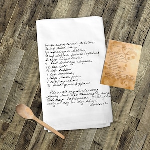 May include: A white kitchen towel with handwritten instructions for a recipe called "Broccoli Salad". The recipe includes ingredients like cubed corn, diced celery, chopped broccoli, chopped pimento, hard-boiled eggs, mayonnaise, lemon juice, and green peppers. The towel is on a wooden surface with a wooden spoon.