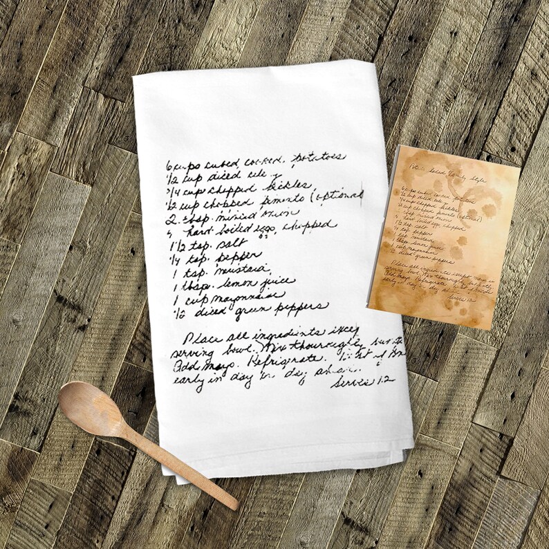 May include: A white kitchen towel with handwritten instructions for a recipe called "Broccoli Salad". The recipe includes ingredients like cubed corn, diced celery, chopped broccoli, chopped pimento, hard-boiled eggs, mayonnaise, lemon juice, and green peppers. The towel is on a wooden surface with a wooden spoon.