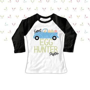 May include: A white and black raglan-style shirt with the text "Expert Egg Hunter Payton". The shirt features a light blue truck carrying Easter eggs and a bunny. The background has a pattern of pastel bunny ears.