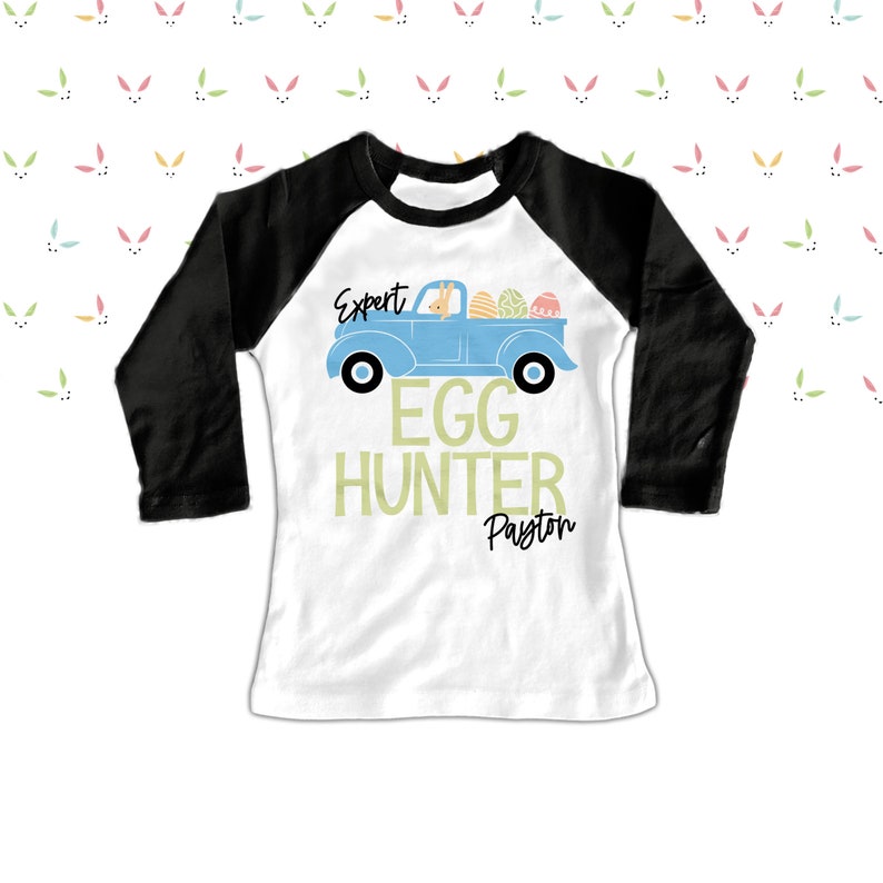 May include: A white and black raglan-style shirt with the text "Expert Egg Hunter Payton". The shirt features a light blue truck carrying Easter eggs and a bunny. The background has a pattern of pastel bunny ears.