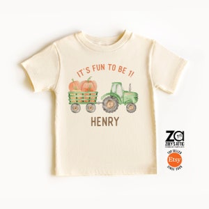 May include: A beige short-sleeved t-shirt with a green tractor pulling a wagon full of orange pumpkins. The text "IT'S FUN TO BE 1!" is above the tractor and the name "HENRY" is below the tractor.