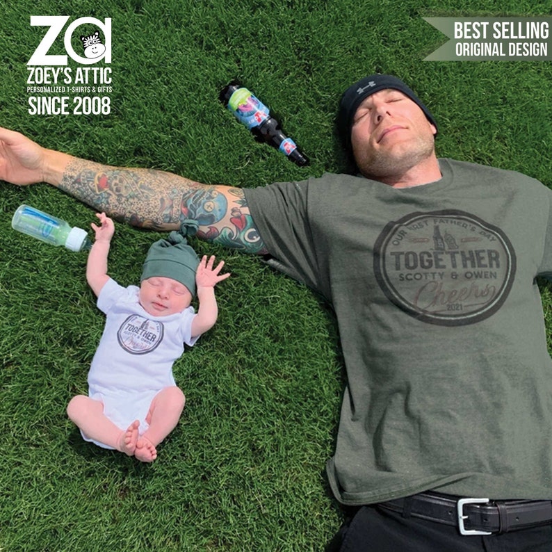 May include: A father and son wearing matching green t-shirts with a white circle graphic that says "Our Most Father's Day Together Scotty & Owen Cheers 2021". The father is lying on his back on a green grassy lawn. The son is lying on his back on the grass with his arms outstretched.