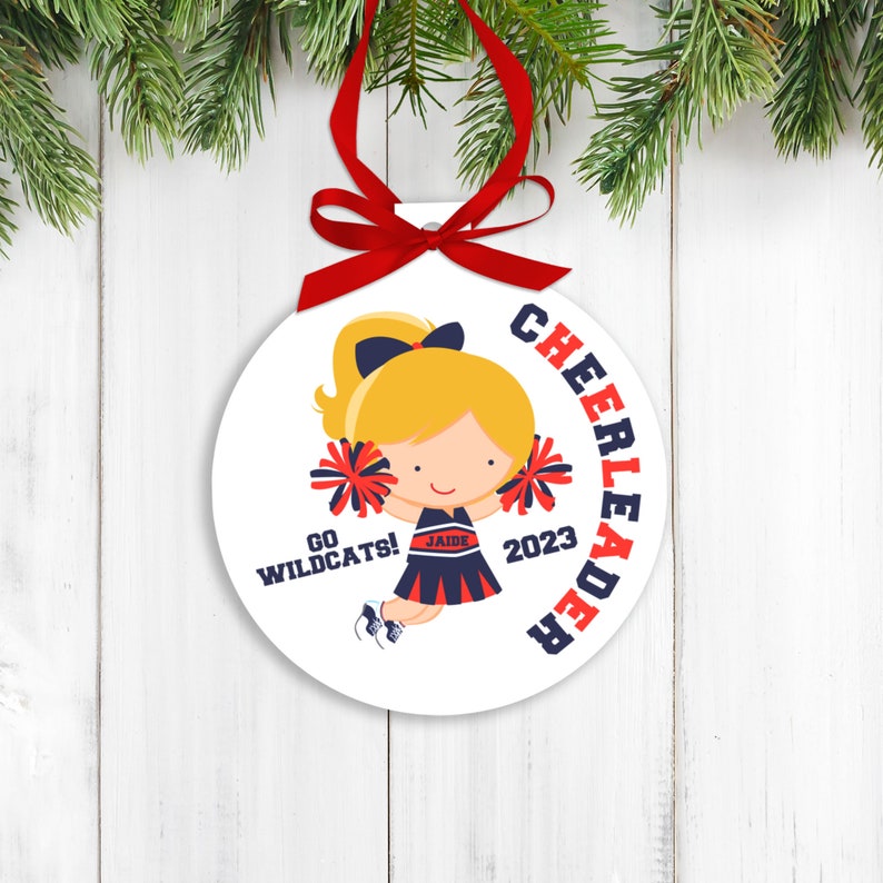 May include: Round white ornament with a cartoon cheerleader in a red, white, and blue uniform. The ornament says "Cheerleader" in a red circle around the cheerleader, "Go Wildcats!" at the bottom left, "2023" at the bottom right, and "Jaide" on the cheerleader's uniform.