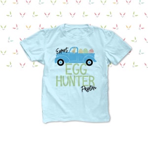 May include: A pale blue t-shirt with a graphic of a blue truck filled with Easter eggs. The text on the shirt says "Expert Egg Hunter Payton". The background is white with a pattern of bunny ears.
