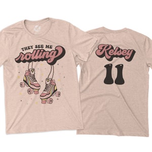 Puede incluir: Una camiseta rosa claro con el texto "They See Me Rolling" en una fuente retro con un coraz&oacute;n en la palabra "rolling". La camiseta presenta un par de patines rosas con rayas arco&iacute;ris y un coraz&oacute;n en cada pat&iacute;n. La parte posterior de la camiseta tiene el texto "Kelsey 11" en una fuente retro.