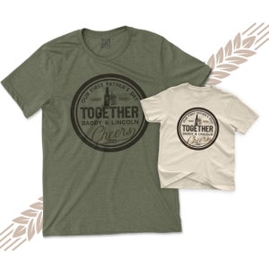 May include: Two green and beige t-shirts with a brown circle graphic that says "Our First Father's Day Together Daddy & Lincoln Cheers 2023".