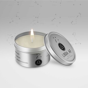 May include: A silver-colored candle tin with a lit wick and a lid. The lid is slightly ajar, revealing the candle inside. The lid has the text "LIBRA AF" and a Libra symbol. The candle is labeled with the words "baffling", "flirting", and "people-pleasing".