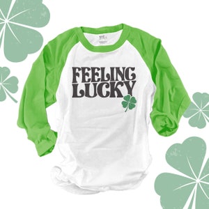 May include: White and green raglan shirt with the text "FEELING LUCKY" in black lettering and a green shamrock graphic. The shirt has green sleeves and a white body, perfect for St. Patrick's Day.