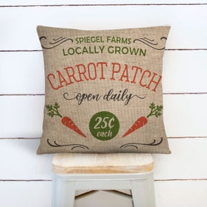 May include: A brown burlap pillow with the text "Spiegel Farms Locally Grown Carrot Patch open daily 25c each" in green, orange, and white lettering. The pillow features two orange carrots with green tops.