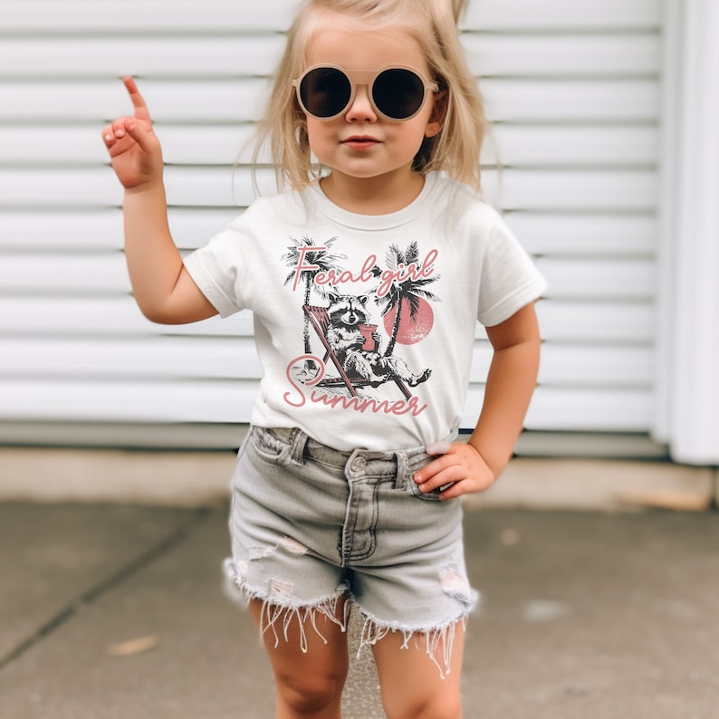 May include: A white t-shirt with a graphic of a raccoon sitting in a beach chair with a drink. The text "Feral girl summer" is printed in pink on the shirt.