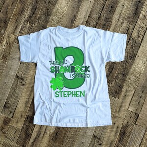May include: White t-shirt with a green shamrock design and the text "This Lil' Shamrock is Three!" with a name "Stephen" underneath.
