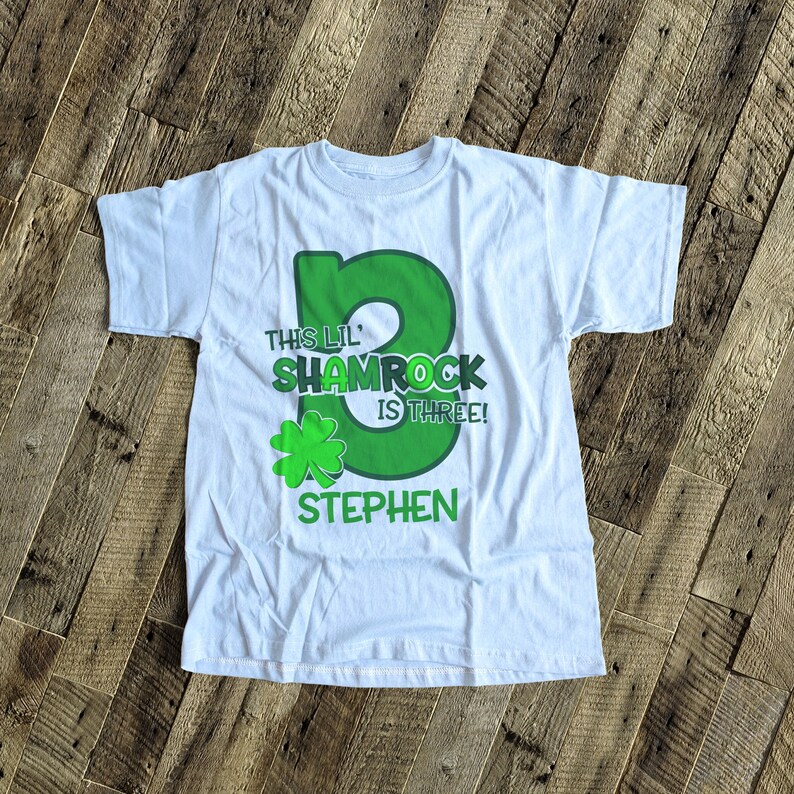 May include: White t-shirt with a green shamrock design and the text "This Lil' Shamrock is Three!" with a name "Stephen" underneath.