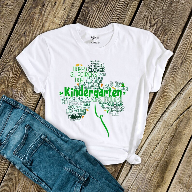 以下が含まれることがあります： 緑のクローバーのデザインが施された白いTシャツで、「Kindergarten」と「Mrs. Pollard」の文字が書かれています。デザインには、「クローバー」、「シャムロック」、「妖精」、「虹」など、聖パトリックの日に関連する他の単語も含まれています。