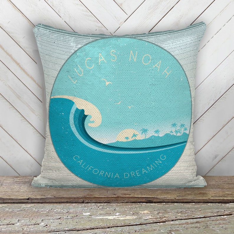 May include: A turquoise blue and white pillow with a circular design featuring a wave, palm trees, and the text "Lucas Noah California Dreaming".