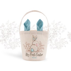 May include: A light beige canvas Easter basket with blue bunny ears. The basket features a cute bunny illustration and the text "My First Easter Wyatt".
