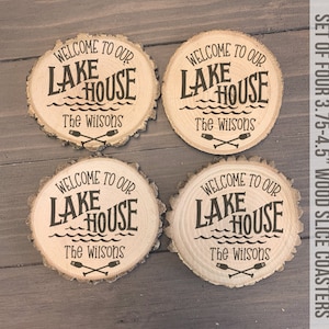 May include: Set of four 3.75-4.5 inch wood slice coasters with the text "Welcome to our Lake House The Wilsons" and a graphic of crossed oars and waves.