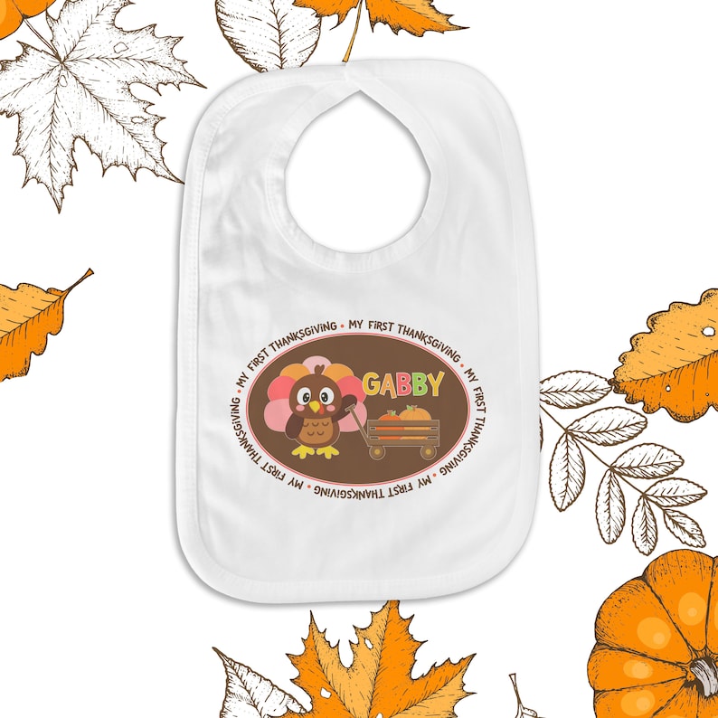 May include: White baby bib with a brown oval design featuring a cartoon turkey with the text "My First Thanksgiving" repeated around the border and the name "Gabby" in pink letters.