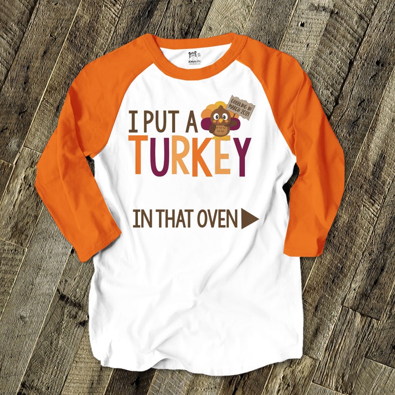Pu&ograve; includere: Maglietta bianca e arancione in stile raglan con la scritta "I PUT A TURKEY IN THAT OVEN" in marrone, arancione e viola. Un tacchino dei cartoni animati &egrave; raffigurato sopra la parola "oven". La maglietta ha anche un piccolo cartello con la scritta "Gobbling in March 2021".