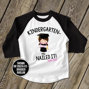 May include: White and black baseball style t-shirt with the text "Kindergarten- Nailed It! 2018-2019" and a cartoon image of a girl wearing a graduation cap and gown holding a diploma. The text "Claire" is on the diploma.