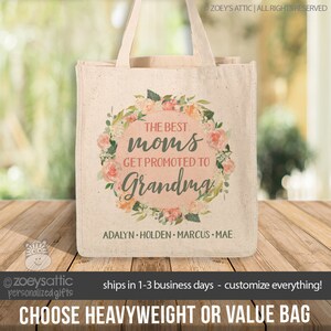 May include: A white canvas tote bag with a floral wreath design. The wreath has pink and peach flowers and green leaves. The text in the center reads "The Best Moms Get Promoted to Grandma". Below the wreath, the names "Adalyn, Holden, Marcus, Mae" are printed.