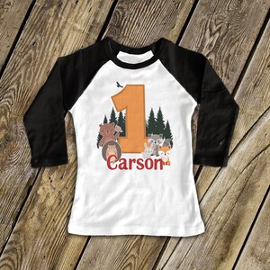 May include: White and black baseball tee shirt with an orange number one and woodland animals. The shirt says "Carson" in red.