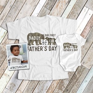 May include: White t-shirt with a brown bear silhouette and the text "Daddy Our First Cub 20 20 Father's Day". A smaller white t-shirt with the same design is shown next to the larger t-shirt. The smaller t-shirt has the text "#FirstDadsDay" printed below it.