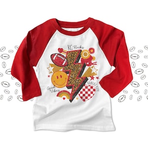 May include: White and red long-sleeved baseball tee with a graphic design featuring a lightning bolt, a smiley face, a football, a chequered heart, and the text "KC Rocks" and "Kansas City".