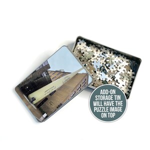 May include: A metal tin with a jigsaw puzzle inside. The tin has a picture of a large baseball bat leaning against a building. The text on the bat reads "Louisville Slugger". The text on the tin reads "Add-on storage tin will have the puzzle image on top."
