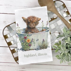 May include: A watercolor illustration of a brown Highland cow taking a bath in a white tub with flowers and greenery around it. The text "highland, illinois" is printed below the tub.