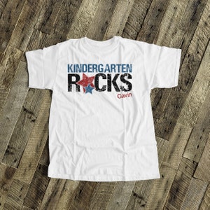 May include: White t-shirt with the text "Kindergarten Rocks" in black with red and blue stars. The name "Gavin" is printed below the text.