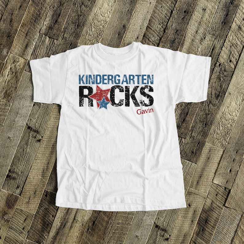 May include: White t-shirt with the text "Kindergarten Rocks" in black with red and blue stars. The name "Gavin" is printed below the text.