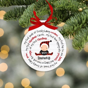 May include: A white round Christmas ornament with a red ribbon. The ornament features a baby in a red and black plaid outfit with the text "my first smell of freshly baking cookies... my first Christmas carols... my first time ripping into presents... my first trip to see Santa" and "2023 Tommy".
