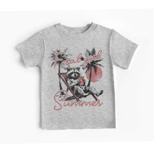 May include: Gray t-shirt with a raccoon wearing sunglasses and holding a drink, sitting in a beach chair under palm trees. The text "Feral girl summer" is printed on the shirt.