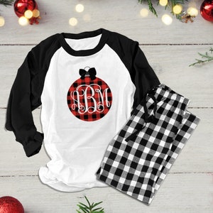 Op de afbeelding: Witte en zwarte geruite pyjamaset met een shirt met lange mouwen met een rood en zwart buffalo plaid ornament met een zwarte strik en een wit monogram "BM".