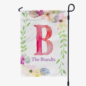 May include: A white garden flag with a pink watercolor letter B surrounded by a wreath of green leaves, watercolor flowers, and speckled eggs. The text "The Brandts" is written in purple below the letter.