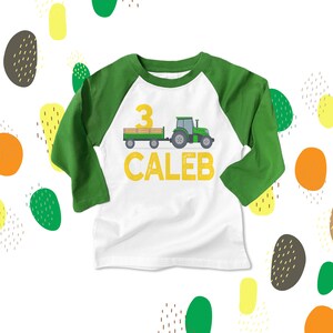 Puede incluir: Una camiseta de b&eacute;isbol de manga larga blanca y verde con un tractor verde y un remolque, el n&uacute;mero 3 y el nombre Caleb en amarillo.