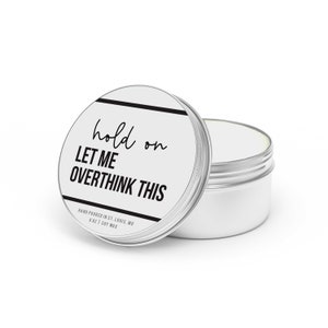 May include: A silver candle tin with a white label that reads "hold on LET ME OVERTHINK THIS" and "HAND POURED IN ST. LOUIS, MO 6 OZ | SOY WAX".