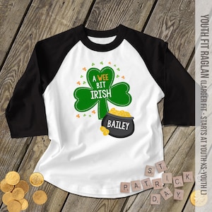May include: A white and black baseball tee shirt with a green shamrock and the text "A Wee Bit Irish" and "Bailey". The shirt is decorated with gold coins.