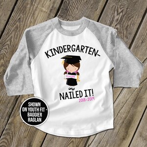 May include: White and grey youth baseball t-shirt with the text "Kindergarten- Nailed It! 2018-2019" and a cartoon illustration of a girl wearing a graduation cap and gown holding a scroll that says "Claire".
